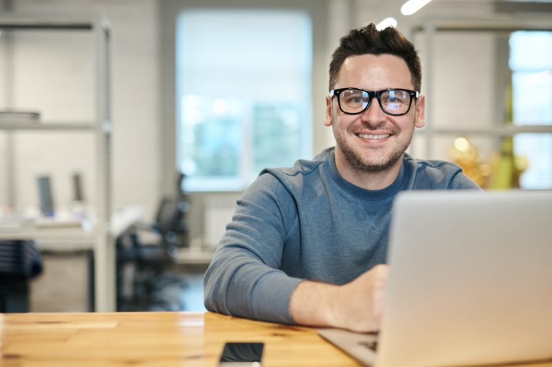 パソコンをしながらこちらをむいて笑顔のメガネをかけた男性