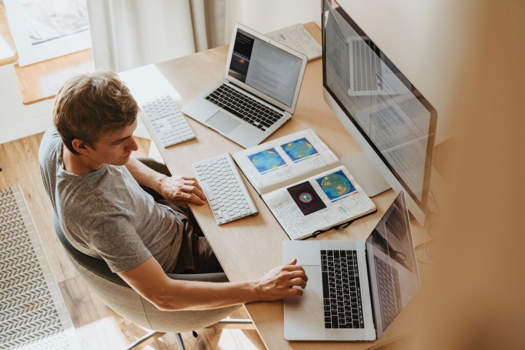 パソコンを3台机に並べて操作するグレーのTシャツの男性