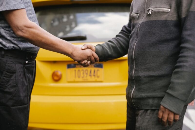 黄色い車の前でパジャマのような格好で握手する二人の男性